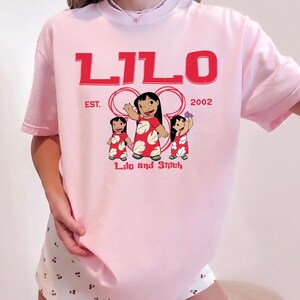 Puede incluir: Una camiseta de manga corta de color rosa claro con un estampado gr&aacute;fico rojo de Lilo de Lilo y Stitch. El texto dice LILO, EST. 2002 y Lilo and Stitch. El dise&ntilde;o incluye tres ilustraciones del personaje Lilo con un vestido rojo de flores.