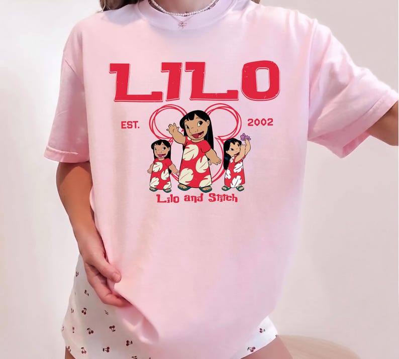 Puede incluir: Una camiseta de manga corta de color rosa claro con un estampado gr&aacute;fico rojo de Lilo de Lilo y Stitch. El texto dice LILO, EST. 2002 y Lilo and Stitch. El dise&ntilde;o incluye tres ilustraciones del personaje Lilo con un vestido rojo de flores.