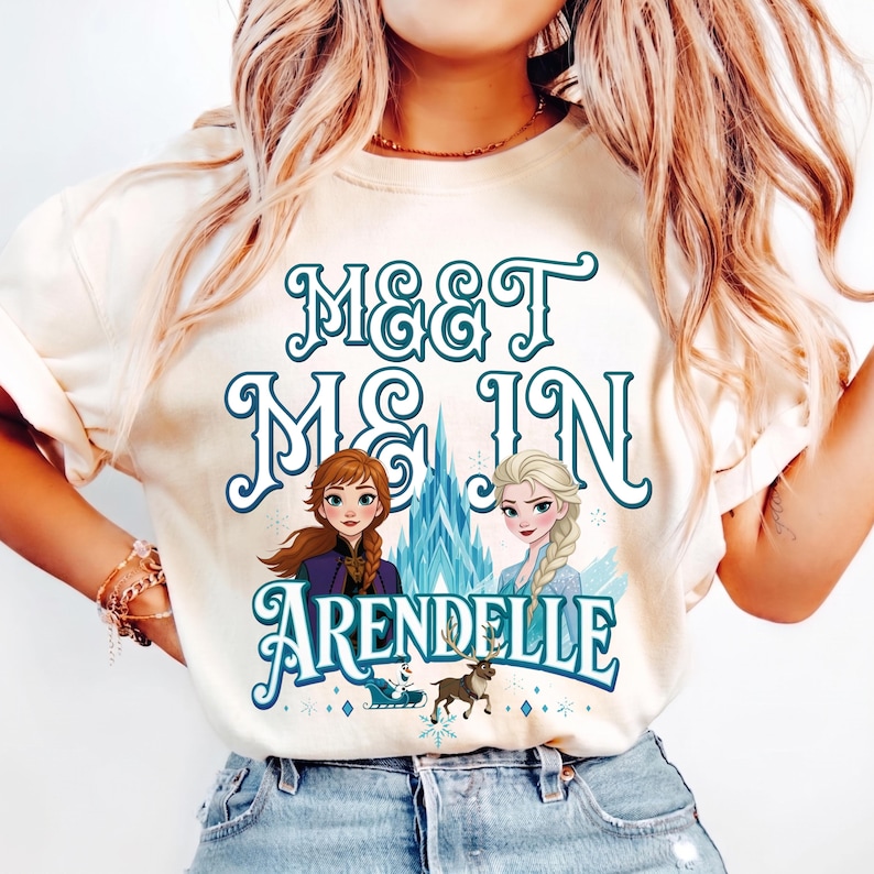 May include: Cream-colored t-shirt with the text "MEET ME IN ARENDELLE" in teal script. The graphic features illustrations of two princesses, a castle, and a reindeer. The shirt is made of a soft material.