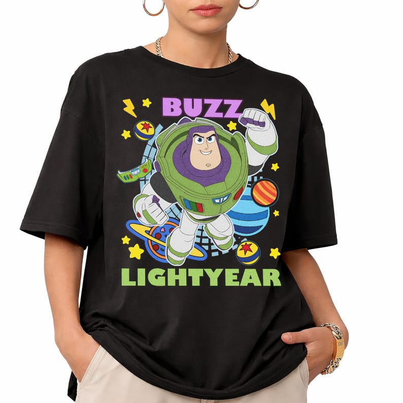 May include: Black t-shirt featuring a colorful graphic of Buzz Lightyear from Toy Story. The design includes the words "BUZZ" and "LIGHTYEAR" in purple and green, with stars and planets. The shirt has short sleeves and a crew neck.