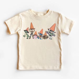 May include: Cream-colored t-shirt featuring a graphic of cartoon characters wearing orange traffic cones. The characters include a green dinosaur, a cowboy, a pink pig, a blue alien, a brown dog, and a green space ranger.