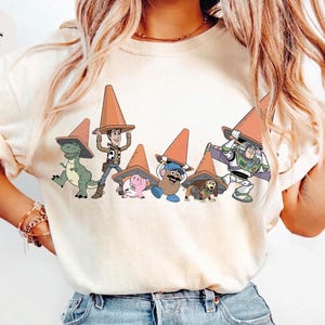 May include: Ivory-colored t-shirt featuring a graphic of Toy Story characters wearing orange traffic cones. The characters include Rex, Woody, Hamm, Mr. Potato Head, Slinky Dog, and Buzz Lightyear. The text "Colors Ivory" is in the upper left corner.