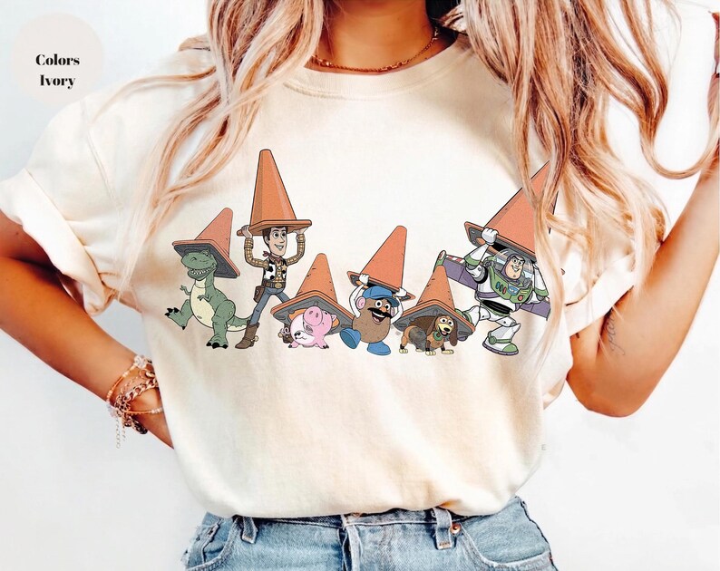May include: Ivory-colored t-shirt featuring a graphic of Toy Story characters wearing orange traffic cones. The characters include Rex, Woody, Hamm, Mr. Potato Head, Slinky Dog, and Buzz Lightyear. The text "Colors Ivory" is in the upper left corner.