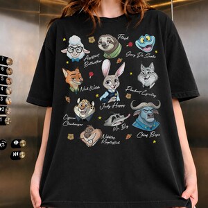 May include: A black t-shirt featuring a graphic print of various animated animal characters from Zootopia, including Judy Hopps, Nick Wilde, Flash, and Chief Bogo, with their names printed in white script text alongside small yellow stars and carrots.