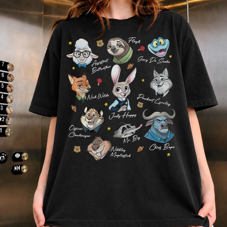 May include: A black t-shirt featuring a graphic print of various animated animal characters from Zootopia, including Judy Hopps, Nick Wilde, Flash, and Chief Bogo, with their names printed in white script text alongside small yellow stars and carrots.