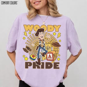 May include: A lavender Comfort Colors t-shirt featuring a graphic of Woody from Toy Story. The design includes the word "WOODY" in yellow, Woody's image, and the word "PRIDE" in brown. Additional graphics include stars, a hat, and a block with the letter "A".