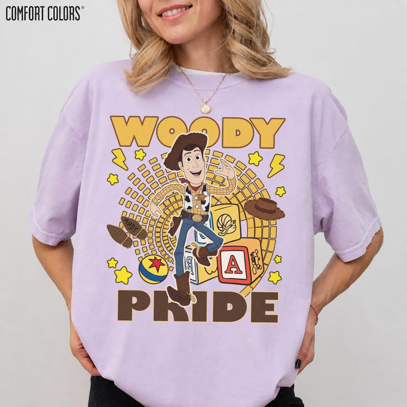 May include: A lavender Comfort Colors t-shirt featuring a graphic of Woody from Toy Story. The design includes the word "WOODY" in yellow, Woody's image, and the word "PRIDE" in brown. Additional graphics include stars, a hat, and a block with the letter "A".