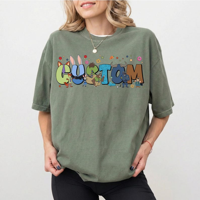 Pode incluir: Uma t-shirt de manga curta verde-azeitona com a palavra CUSTOM em letras grandes, coloridas e em estilo bloco. Cada letra incorpora v&aacute;rias personagens de desenhos animados e pequenas formas decorativas como estrelas, flores e donuts.