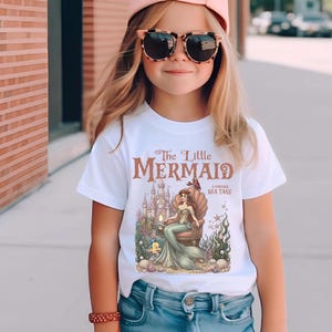 May include: White t-shirt featuring a vintage-style graphic of a mermaid sitting on a throne in front of a castle. The text "The Little Mermaid" is displayed above the image. The shirt is worn by a young person.