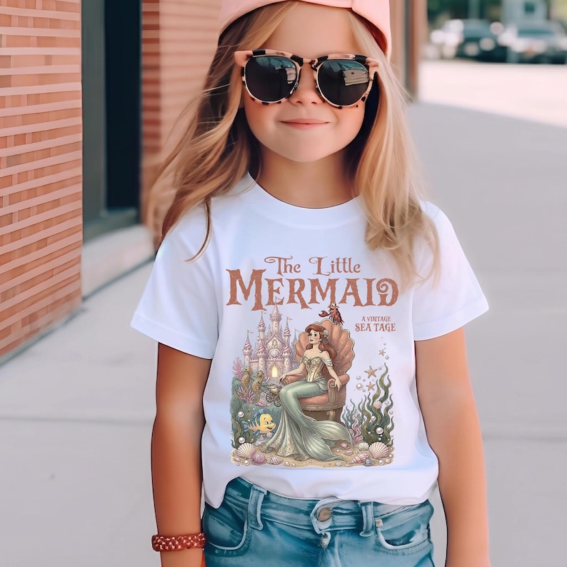 May include: White t-shirt featuring a vintage-style graphic of a mermaid sitting on a throne in front of a castle. The text "The Little Mermaid" is displayed above the image. The shirt is worn by a young person.