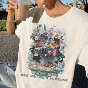 May include: Cream-colored t-shirt featuring a colorful graphic of animated animal characters. The design includes the text "We're wild and we can't be tamed" in a rainbow font. The shirt is made of a soft material.