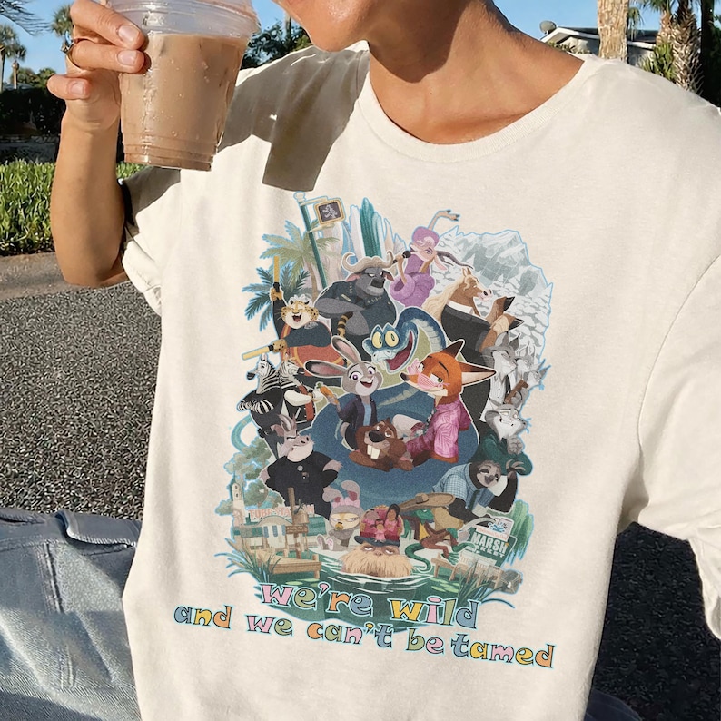 May include: Cream-colored t-shirt featuring a colorful graphic of animated animal characters. The design includes the text "We're wild and we can't be tamed" in a rainbow font. The shirt is made of a soft material.