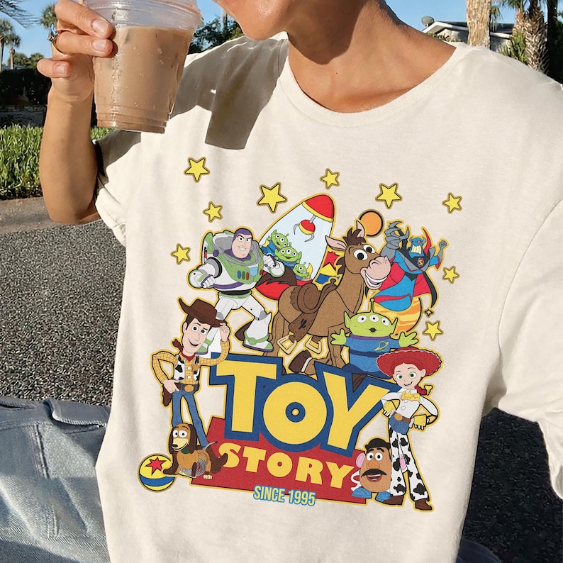 May include: Cream-colored t-shirt featuring a colorful graphic of characters from the animated film "Toy Story." The design includes Buzz Lightyear, Woody, and other characters, with the words "TOY STORY" and "SINCE 1995."