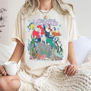 May include: Cream-colored t-shirt featuring a graphic of Ariel from The Little Mermaid. The design includes Ariel, Flounder, and Sebastian, with the words "The Little Mermaid" in a purple font. The shirt has a relaxed fit.