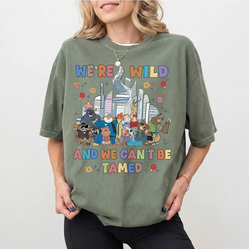 K&ouml;nnte beinhalten: Ein olivgr&uuml;nes Kurzarm-T-Shirt mit einer bunten Grafik verschiedener animierter Tierfiguren vor einer Stadtsilhouette. Der Text "WE'RE WILD" und "AND WE CAN'T BE TAMED" ist in mehrfarbigen Buchstaben &uuml;ber und unter den Figuren aufgedruckt.