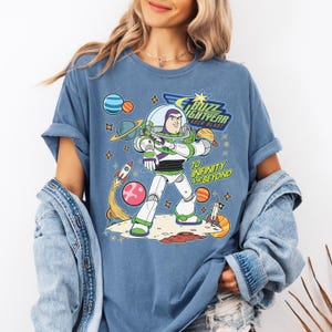 May include: Blue t-shirt featuring Buzz Lightyear from Toy Story. The graphic includes the text "Buzz Lightyear Laser Blast" and "To Infinity and Beyond" with planets, rockets, and stars. The shirt has rolled-up sleeves.