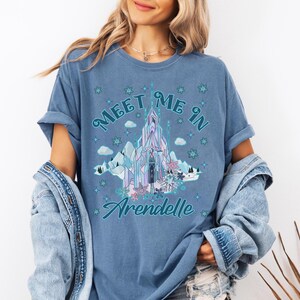 May include: A blue t-shirt with the text "Meet Me In Arendelle" and a graphic of a castle, mountains, and snowflakes. The design is in shades of blue, purple, and white. The shirt has rolled-up sleeves.