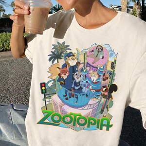 Puede incluir: Camiseta color crema con un gr&aacute;fico colorido de la pel&iacute;cula "Zootopia". El dise&ntilde;o incluye varios personajes animales, el horizonte de la ciudad y la palabra "Zootopia" en una fuente verde y azul vibrante. La camiseta es de material suave.