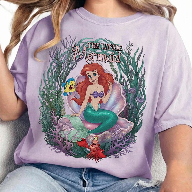 May include: A light purple t-shirt featuring a graphic of Ariel from The Little Mermaid. Ariel, with red hair and a green tail, sits in a seashell. The shirt includes the text "THE LITTLE Mermaid" and images of Flounder and Sebastian.