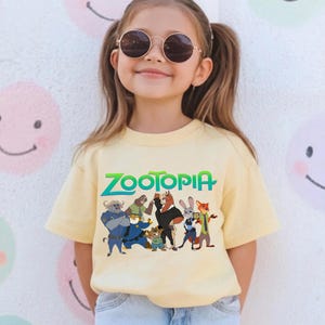 K&ouml;nnte beinhalten: Ein hellgelbes T-Shirt mit dem Wort "Zootopia" in Gr&uuml;n und Blau und Zeichentrickfiguren aus dem Film. Das T-Shirt wird von einer jungen Person getragen, die eine runde Sonnenbrille tr&auml;gt.