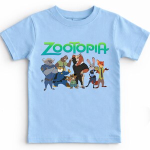 K&ouml;nnte beinhalten: Ein hellblaues T-Shirt mit dem Wort "ZOOTOPIA" in Gr&uuml;n und einer Gruppe von Zeichentrickfiguren. Die Figuren umfassen einen Polizisten, ein Faultier, einen Tiger, ein Pferd, ein Kaninchen und einen Fuchs.