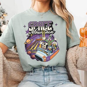 May include: A pale green t-shirt with a vibrant graphic of the Space Mountain ride, showcasing Mickey Mouse, Donald Duck, Goofy, and Pluto. The words "SPACE MOUNTAIN" are printed above the ride scene.
