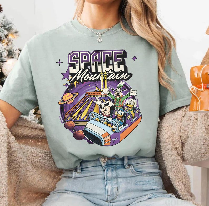 May include: A pale green t-shirt with a vibrant graphic of the Space Mountain ride, showcasing Mickey Mouse, Donald Duck, Goofy, and Pluto. The words "SPACE MOUNTAIN" are printed above the ride scene.
