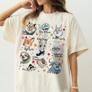 May include: A cream-colored t-shirt featuring a graphic print of various animated animal characters from Zootopia, including Judy Hopps, Nick Wilde, Flash, and Chief Bogo, each labeled with their respective names in a black cursive script font.