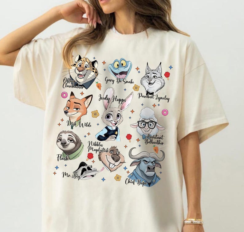 May include: A cream-colored t-shirt featuring a graphic print of various animated animal characters from Zootopia, including Judy Hopps, Nick Wilde, Flash, and Chief Bogo, each labeled with their respective names in a black cursive script font.