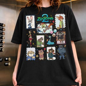 May include: A black short-sleeve t-shirt featuring a graphic print with the text Zootopia 2. The design includes multiple framed illustrations of various animated animal characters from the movie, including a rabbit, fox, sloth, and others.