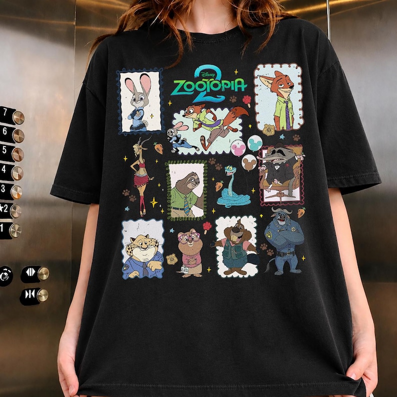 May include: A black short-sleeve t-shirt featuring a graphic print with the text Zootopia 2. The design includes multiple framed illustrations of various animated animal characters from the movie, including a rabbit, fox, sloth, and others.