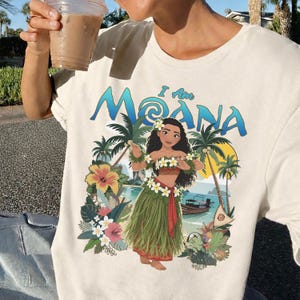 May include: Cream-colored t-shirt featuring a colorful graphic of a woman in a grass skirt, with the text "I Am Moana". The design includes palm trees, flowers, and a boat, evoking a tropical theme.