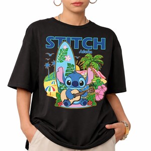 May include: Black t-shirt featuring a colorful graphic of Stitch playing a guitar on a tropical beach. The design includes a surfboard, palm trees, and the text "STITCH Aloha" in blue and white.