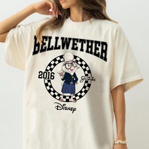May include: An off-white t-shirt featuring a graphic of the character Bellwether from Disney. The design includes the text BELLWETHER, 2016, and Disney, centred within a black and white checkered circle. The character wears glasses and a blue outfit.