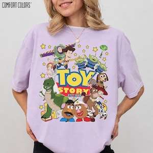 May include: A light purple Comfort Colors t-shirt featuring a colorful graphic of characters from the animated film "Toy Story." The design includes Woody, Buzz Lightyear, the aliens, and other characters, with the text "TOY STORY EST.1995."