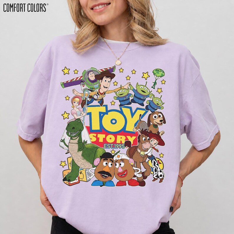 May include: A light purple Comfort Colors t-shirt featuring a colorful graphic of characters from the animated film "Toy Story." The design includes Woody, Buzz Lightyear, the aliens, and other characters, with the text "TOY STORY EST.1995."