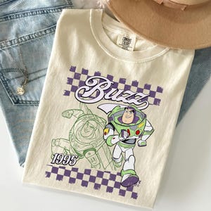 May include: Cream-colored t-shirt featuring a graphic of Buzz Lightyear from Toy Story. The design includes the word "Buzz" in a stylized font, a checkerboard pattern, and the year "1995". The shirt is folded on top of blue denim jeans.