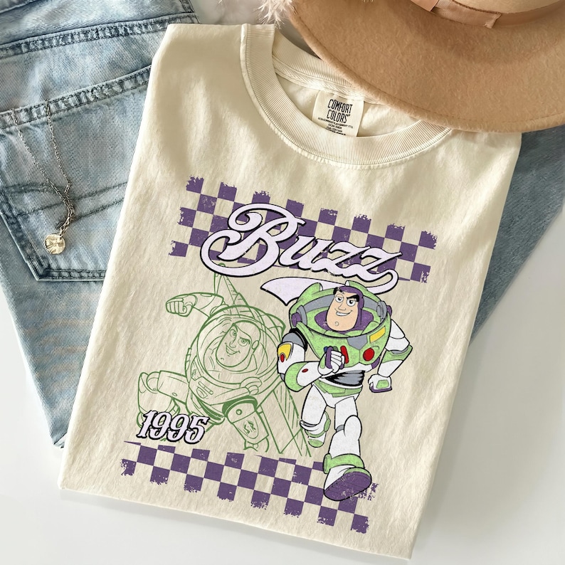 May include: Cream-colored t-shirt featuring a graphic of Buzz Lightyear from Toy Story. The design includes the word "Buzz" in a stylized font, a checkerboard pattern, and the year "1995". The shirt is folded on top of blue denim jeans.