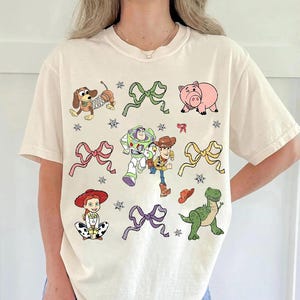 Op de afbeelding: Cr&egrave;mekleurig T-shirt met kleurrijke cartoonfiguren en strikken. Het ontwerp omvat een teckel, een varken, Buzz Lightyear, Woody, Jessie, een dinosaurus en strikken. Het shirt heeft een ronde hals en korte mouwen.