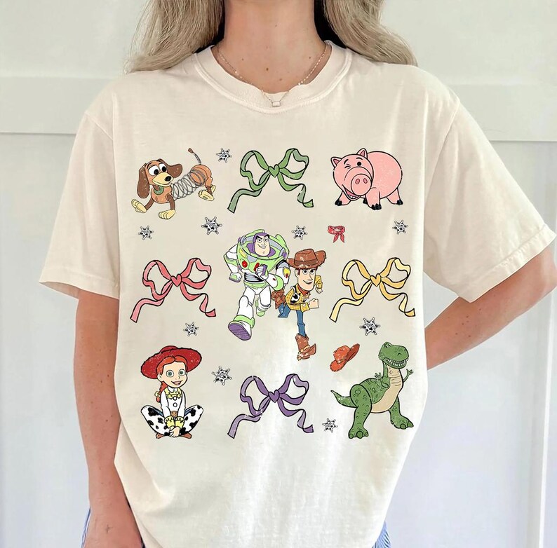 Op de afbeelding: Cr&egrave;mekleurig T-shirt met kleurrijke cartoonfiguren en strikken. Het ontwerp omvat een teckel, een varken, Buzz Lightyear, Woody, Jessie, een dinosaurus en strikken. Het shirt heeft een ronde hals en korte mouwen.