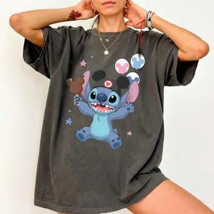 May include: A charcoal gray short-sleeve t-shirt featuring a graphic of the blue character Stitch wearing a black ear hat. Stitch holds a brown chocolate treat and is surrounded by three colorful balloons and small stars on the front chest.