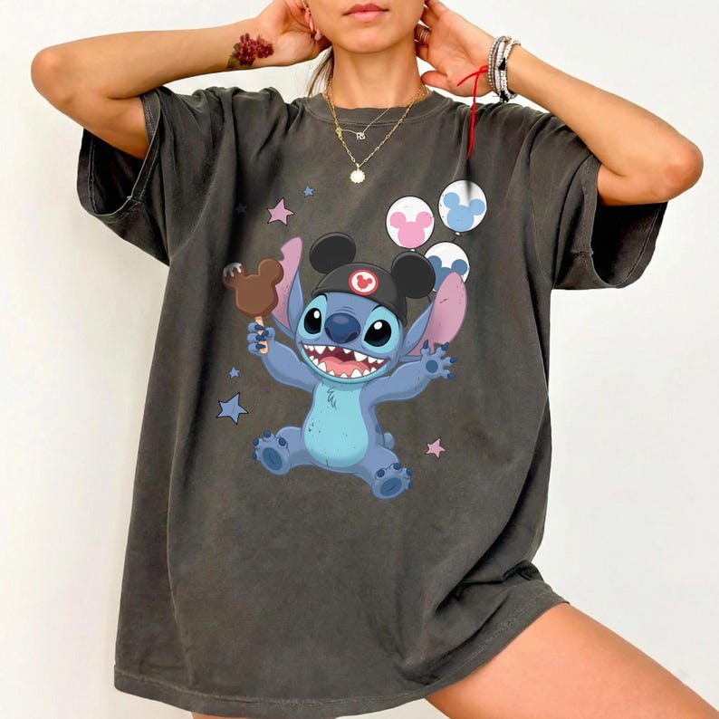 May include: A charcoal gray short-sleeve t-shirt featuring a graphic of the blue character Stitch wearing a black ear hat. Stitch holds a brown chocolate treat and is surrounded by three colorful balloons and small stars on the front chest.