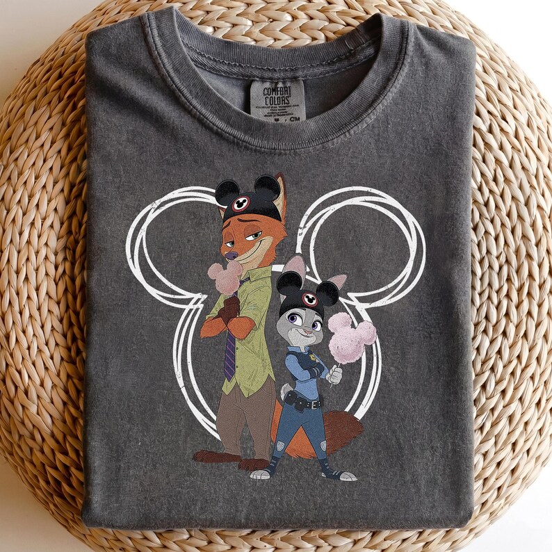 May include: A charcoal gray crew neck t-shirt featuring a graphic of Nick Wilde and Judy Hopps from Zootopia wearing Mickey Mouse ear hats. The characters are positioned in front of a large white outline of Mickey Mouse ears, each holding a pink cotton candy.