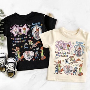 May include: A black t-shirt and a cream t-shirt, each featuring a Zootopia graphic print with characters Judy Hopps and Nick Wilde, floral accents, framed portraits, and the text Welcome to Zootopia, accompanied by a pair of black and white sneakers.