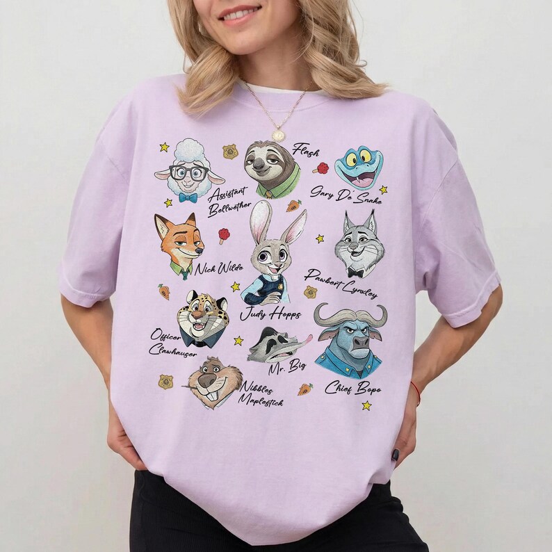 May include: A light purple t-shirt featuring a graphic print of various animated animal characters from Zootopia, including Judy Hopps, Nick Wilde, and Chief Bogo, with their names printed in cursive script alongside small star and carrot icons.