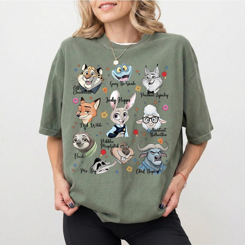 May include: An olive green short-sleeve t-shirt featuring a graphic print of various animated animal characters from Zootopia, including Judy Hopps, Nick Wilde, Flash, and Chief Bogo, with their names printed in black text below each character.