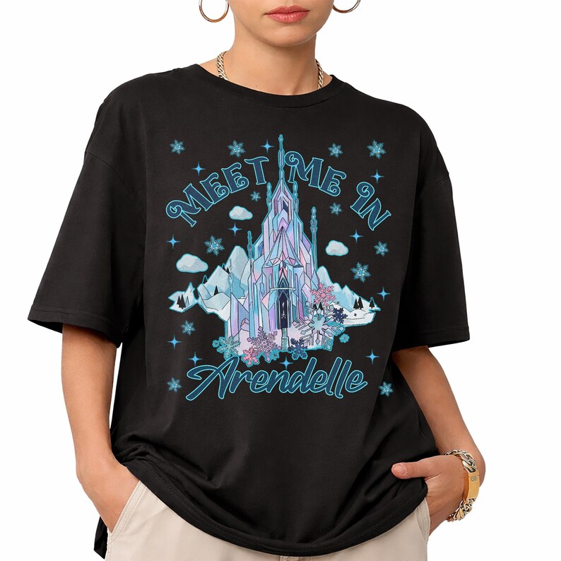 May include: Black t-shirt featuring a graphic of a castle with the text "Meet Me In Arendelle" in blue. The castle design includes shades of blue, purple, and white, with snowflakes and clouds. A casual, oversized fit.