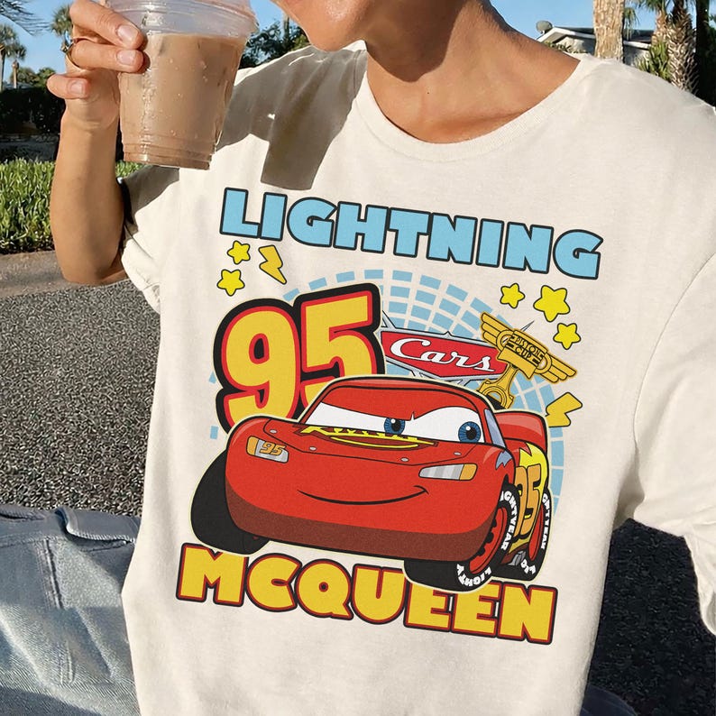 May include: Cream-colored t-shirt featuring a graphic of Lightning McQueen from the movie Cars. The design includes the text "LIGHTNING 95 MCQUEEN" in blue, yellow, and red. The shirt has a crew neck and short sleeves.