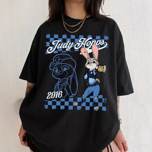 Puede incluir: Una camiseta negra de manga corta con un estampado gr&aacute;fico del personaje Judy Hopps. El dise&ntilde;o incluye el texto "Judy Hopps" y "2016" con un borde a cuadros azules y negros, un uniforme de polic&iacute;a azul y una ilustraci&oacute;n de una placa dorada.
