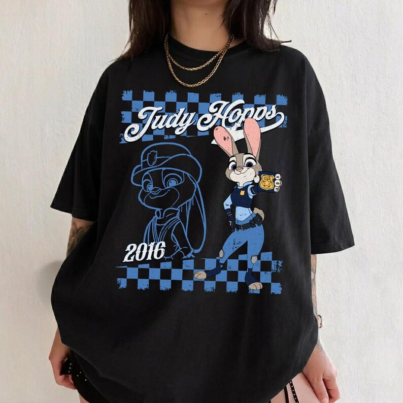 Puede incluir: Una camiseta negra de manga corta con un estampado gr&aacute;fico del personaje Judy Hopps. El dise&ntilde;o incluye el texto "Judy Hopps" y "2016" con un borde a cuadros azules y negros, un uniforme de polic&iacute;a azul y una ilustraci&oacute;n de una placa dorada.